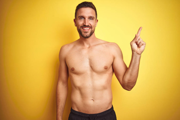 Young handsome shirtless man over isolated yellow background with a big smile on face, pointing with hand and finger to the side looking at the camera. - Fotó, kép