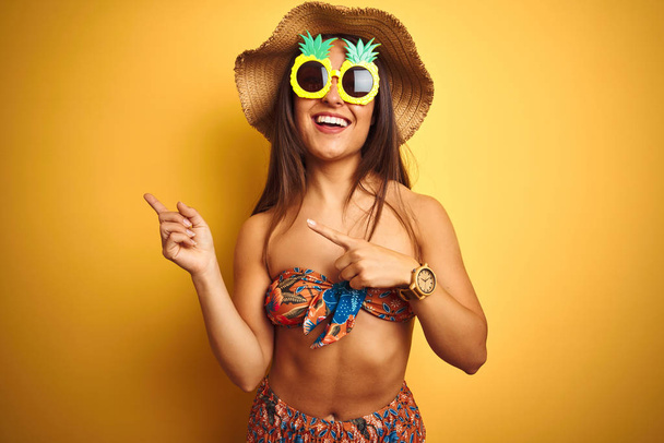 Mujer de vacaciones en bikini y gafas de sol de piña sobre fondo amarillo aislado sonriendo y mirando a la cámara apuntando con dos manos y dedos hacia un lado
. - Foto, imagen