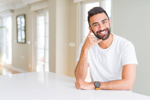Przystojny Hiszpan dorywczo biały t-shirt w domu uśmiechnięty robi gest telefonu z ręki i palców jak rozmawiać przez telefon. Komunikowanie koncepcji. - Zdjęcie, obraz