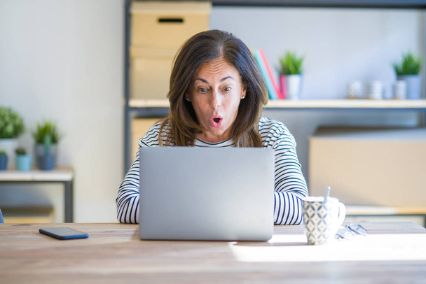 コンピュータラップトップを使用して自宅で仕事をして自宅のテーブルに座っている中年のシニア女性は、驚きの表情、恐怖と興奮した顔でショックを受けました. - 写真・画像