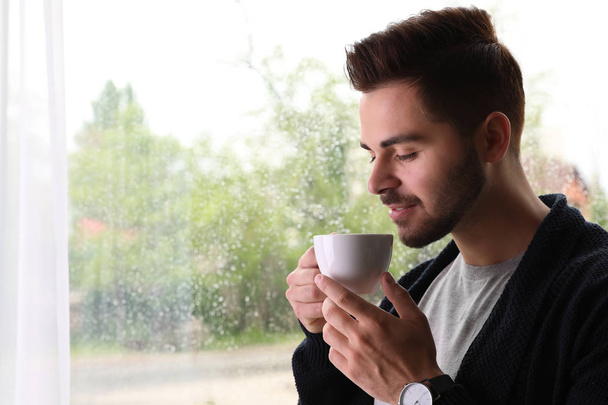 Happy handsome man with cup of coffee near window on rainy day - Foto, imagen