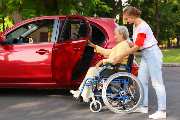 Young woman helping disabled grandmother in wheelchair to get into car outdoors - Foto, Imagen