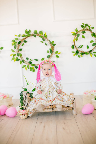 Cute little girl in a dress with a flower print and a bunny hat in the Easter decorations in the studio. Little girl with easter eggs and flowers in a spring studio - Fotografie, Obrázek