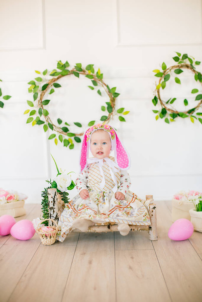 Cute little girl in a dress with a flower print and a bunny hat in the Easter decorations in the studio. Little girl with easter eggs and flowers in a spring studio - Foto, imagen