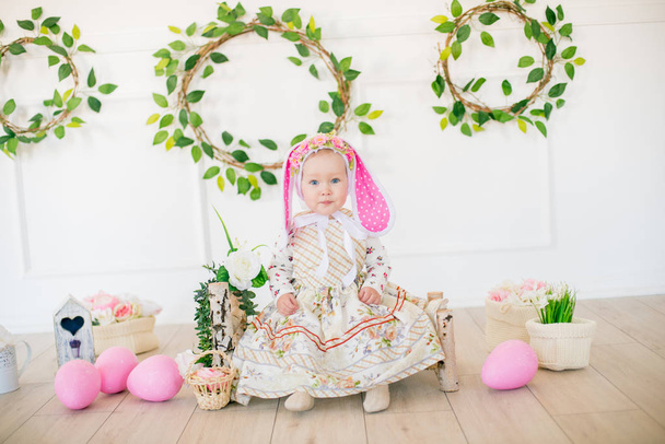 Cute little girl in a dress with a flower print and a bunny hat in the Easter decorations in the studio. Little girl with easter eggs and flowers in a spring studio - Foto, immagini
