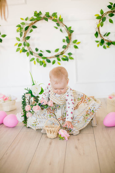 Cute little girl in a dress with a flower print in the Easter decorations in the studio. Little girl with easter eggs and flowers in a spring studio - Foto, immagini