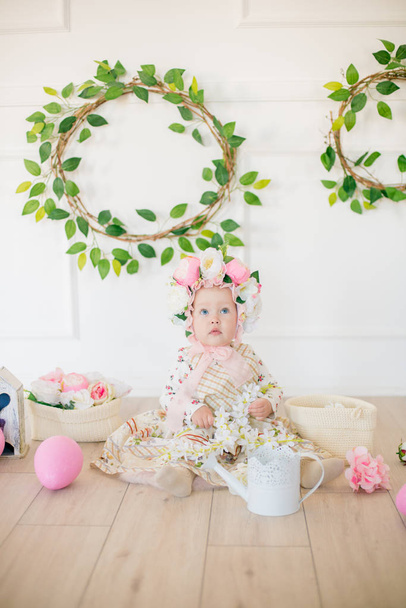 Cute little girl in a dress with a flower print and a hat with flowers in the Easter decorations in the studio. Little girl with easter eggs and flowers in a spring studio - Foto, Imagem