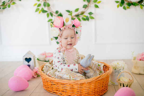 Cute little girl in a dress with a flower print and a hat with flowers in the Easter decorations in the studio. Little girl with easter eggs and flowers in a spring studio - Foto, Imagen