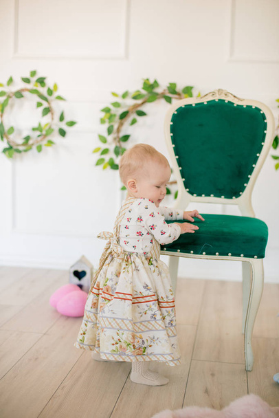 Cute little girl in a dress with a flower print in the Easter decorations in the studio. Little girl with easter eggs and flowers in a spring studio - Photo, Image