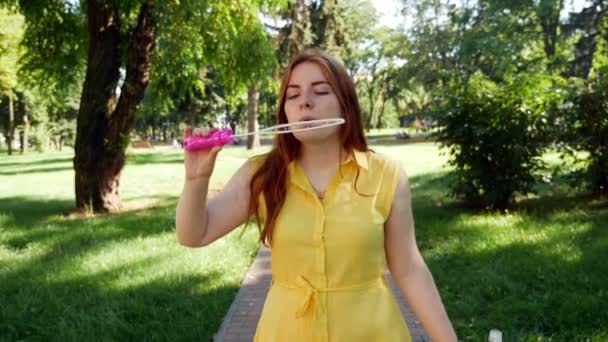 Κοκκινομάλλα κορίτσι χτυπήματα σαπούνι στο πάρκο. κίτρινο φόρεμα χαμογελάει και γελάει. καλοκαίρι και ευτυχία  - Πλάνα, βίντεο