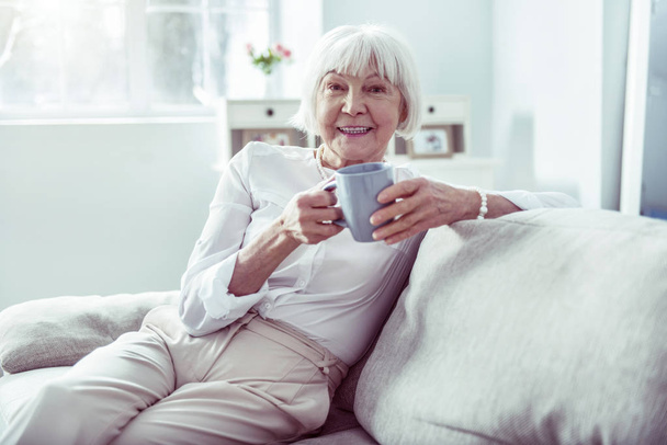 Smiling elderly lady wearing nice stylish white blouse sitting in living room - Foto, afbeelding