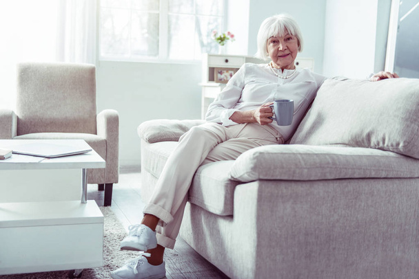 Приємна приємна бабуся сидить на бежевому дивані у вітальні
 - Фото, зображення