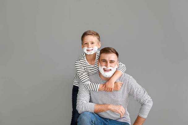 Father and his little son with shaving foam on faces against grey background - Фото, зображення