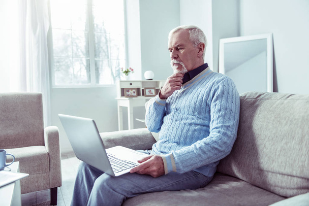 Bearded grey-haired man feeling curious while watching news on laptop - Photo, Image