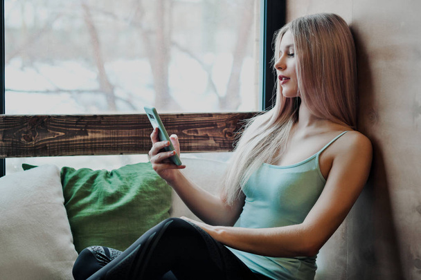 Young beautiful athletic woman blonde in sportswear sitting by the window of a cafe and using a mobile phone. - Foto, Bild