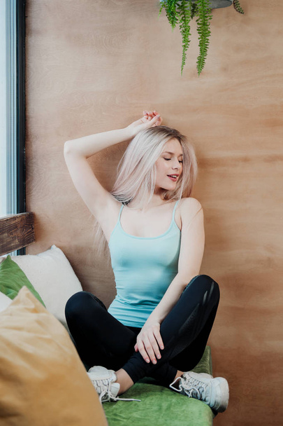 Young beautiful athletic woman blonde in sportswear sitting by the window of a cafe against the background of a wooden wall. - Фото, изображение