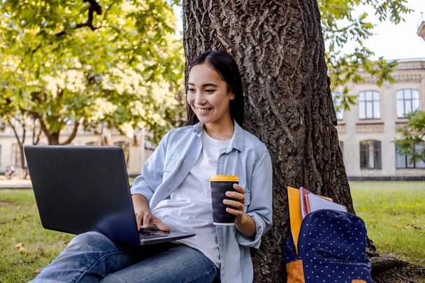 幸せなアジアの学生や女子学生。インターネットを検索し、ノートパソコンを使用し、大学のキャンパスの近くの公園の緑の草の上に座って、試験の準備をします。笑顔、いくつかのニュースを読んで、コーヒーを取り除く飲み物. - 写真・画像
