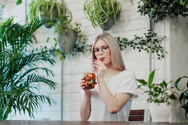 Young beautiful athletic woman blonde in glasses sits at a table in a green cafe and drinks strawberry lemonade from a glass jar. - Photo, Image