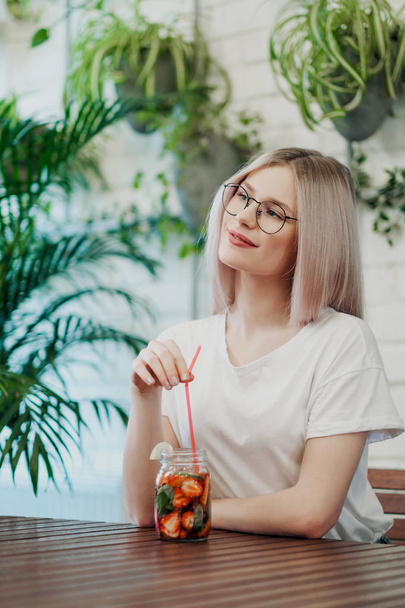Young beautiful athletic woman blonde in glasses sits at a table in a green cafe and drinks strawberry lemonade from a glass jar. - Фото, изображение