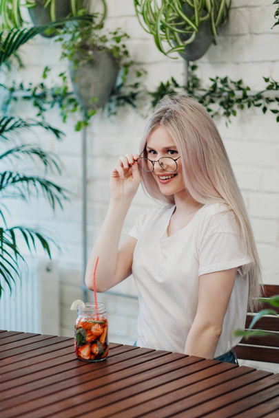 Young beautiful athletic woman blonde in glasses sits at a table in a green cafe and drinks strawberry lemonade from a glass jar. - Фото, изображение