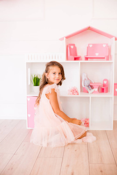 Cute happy girl in a beautiful dress at home plays with a dollhouse and toys - Zdjęcie, obraz