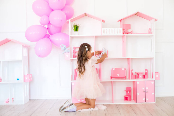 Cute happy girl in a beautiful dress at home plays with a dollhouse and toys - Φωτογραφία, εικόνα