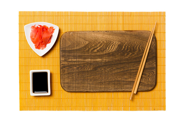 Lege rechthoekige bruine houten plaat met stokjes voor sushi, gember en sojasaus op gele bamboe mat achtergrond. Bovenaanzicht met Kopieer ruimte voor uw ontwerp - Foto, afbeelding