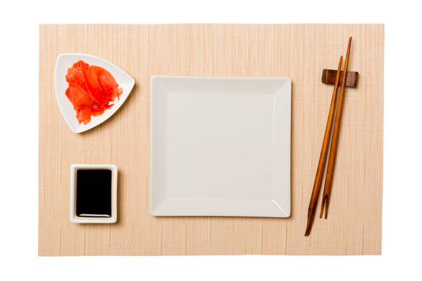 Üres fehér négyzet lemez pálcika a sushi és a szójaszósz, gyömbér a barna sushi szőnyeg háttérben. Felülnézet másolási területtel a tervezéshez - Fotó, kép