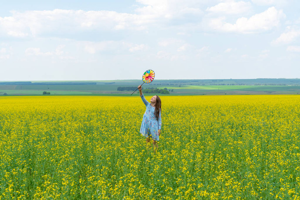 Una muchacha feliz con el pelo largo está parada en la hierba en un campo verde con un molino de viento brillante en sus manos. Concepto de verano, libertad y infancia feliz
 - Foto, imagen