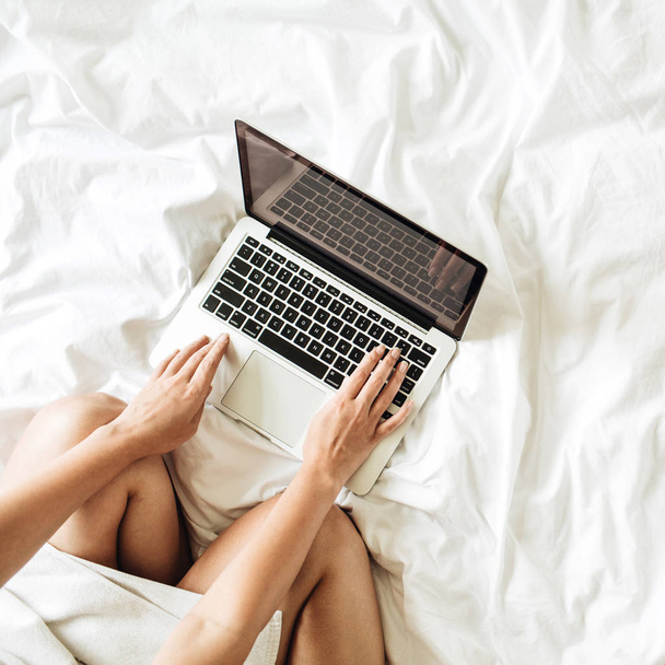 Working at home concept. Girl is working on her laptop in bed with white linens. Flat lay, top view minimalistic workspace background. Relaxing working process.  - Fotografie, Obrázek