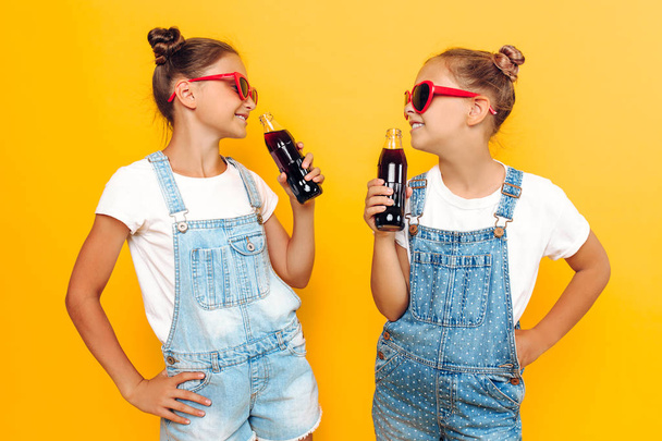 Two teenage girls, girlfriends in sunglasses with soft drinks in their hands, are resting on a yellow background - Foto, imagen