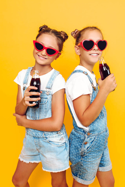 Two teenage girls, girlfriends in sunglasses with soft drinks in their hands, are resting on a yellow background - Fotó, kép