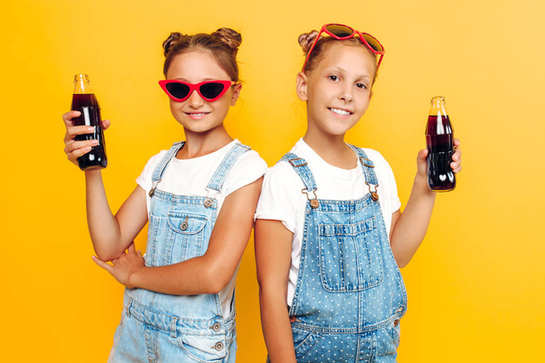 Two teenage girls, girlfriends in sunglasses with soft drinks in their hands, are resting on a yellow background - Foto, Bild
