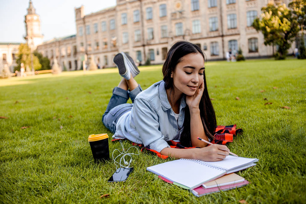 教育コンセプトは、大学キャンパス近くの公園で緑の草を勉強する時間を持つ学生アジアの女性。コーヒーを飲んだり、ノートを書いたり、ノートに書いたりする。学生と女子学生ティーンエイジャーのライフスタイル. - 写真・画像
