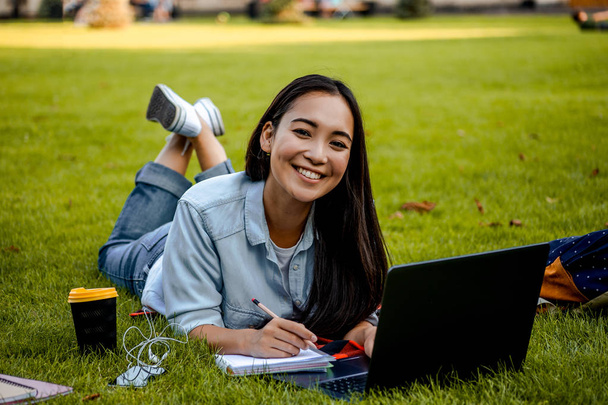 Красивая студентка лежит на траве. Закройте портрет счастливой веселой студентки, делающей домашнее задание, лежа на траве в парке и глядя в камеру. Пользователь портативного компьютера
. - Фото, изображение