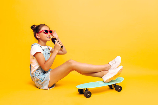 Έφηβος, κορίτσι με γυαλιά ηλίου με ένα σκέιτμπορντ και ένα δροσιστικό ποτό στο χέρι, χαλαρώνοντας σε ένα κίτρινο φόντο - Φωτογραφία, εικόνα