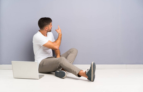 junger Mann mit Laptop, der auf dem Boden sitzt und mit dem Zeigefinger nach hinten zeigt - Foto, Bild