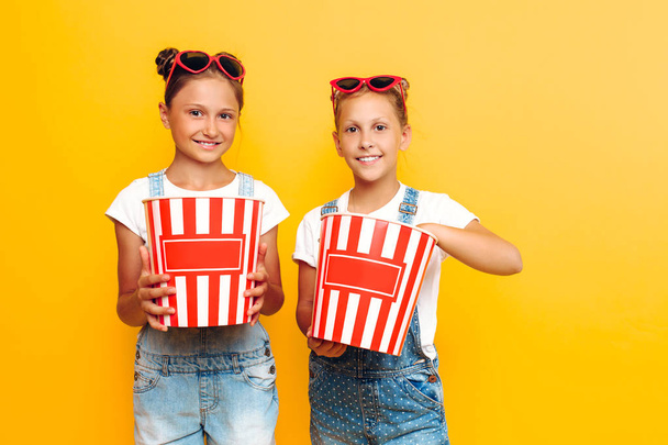 Two teenage girls, girlfriends with popcorn in their hands, posing on a yellow background - Photo, image
