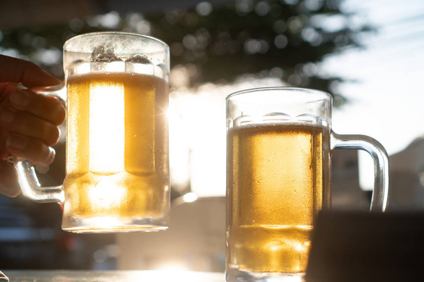 Mão fechada segurando canecas de cerveja lager frescas com raios de sol no dia quente de verão
 - Foto, Imagem