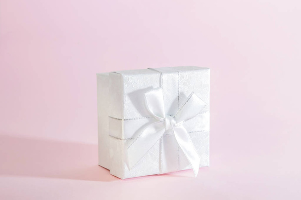 One decorative holiday gift box with a ribbon bow for congratulations, surprise, presentation, white silver on a delicate pink background in pastel colors with a copy space - 写真・画像