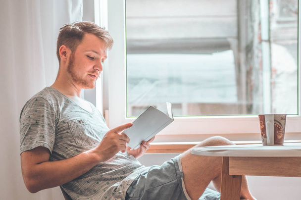 Ελκυστικός νεαρός διαβάζει ένα βιβλίο στο σπίτι. Προνοητικός τύπος που διαβάζει σημαντικό βιβλίο - Φωτογραφία, εικόνα