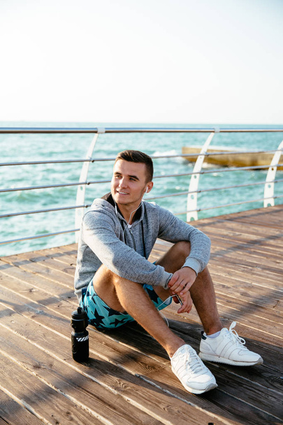 Smiling man in headset, resting after workout, on pier, by the sea. - Photo, image