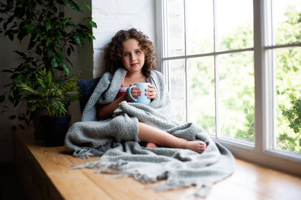 Ένα μικρό κορίτσι κάθεται δίπλα στο παράθυρο με ένα φλιτζάνι τσάι καλυμμένο με μια κουβέρτα. - Φωτογραφία, εικόνα