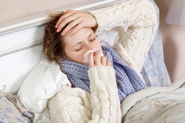 young woman in white sweater and blue scarf cleaning her nose and holding painful head while feeling cold sick and  resting home in bed  - Foto, afbeelding