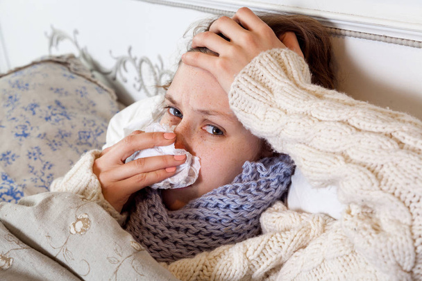 young woman in white sweater and blue scarf cleaning her nose and holding painful head while feeling cold sick and  resting home in bed  - Fotografie, Obrázek