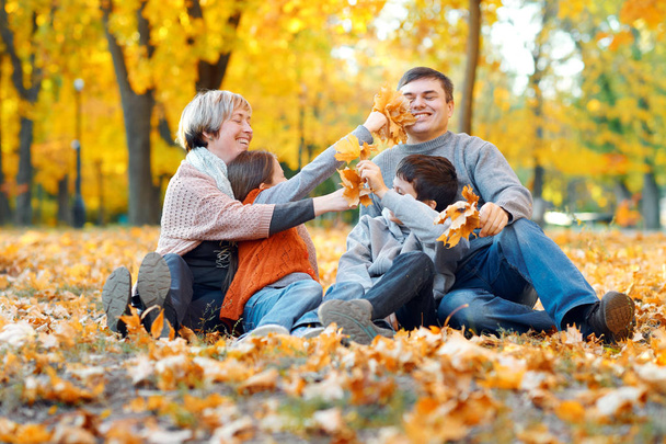 Щаслива сім'я сидить на опале листя, грає і розважається в осінньому міському парку. Діти та батьки разом добре проводять день. Яскраве сонячне світло і жовте листя на деревах, осінній сезон
. - Фото, зображення