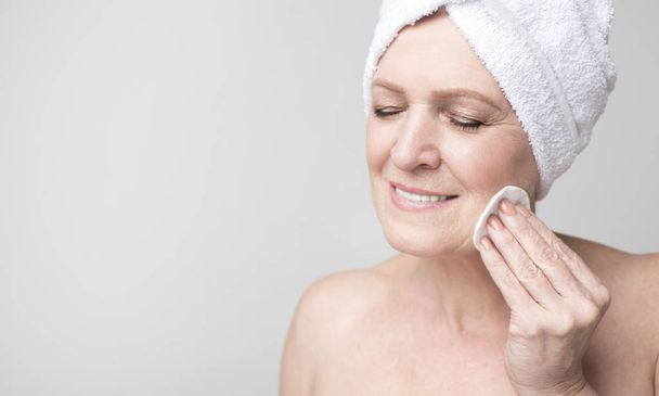 Donna matura con asciugamano sulla testa pulirsi il viso con lozione
 - Foto, immagini