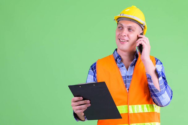 Happy young man construction worker talking on the phone while holding clipboard - Photo, image