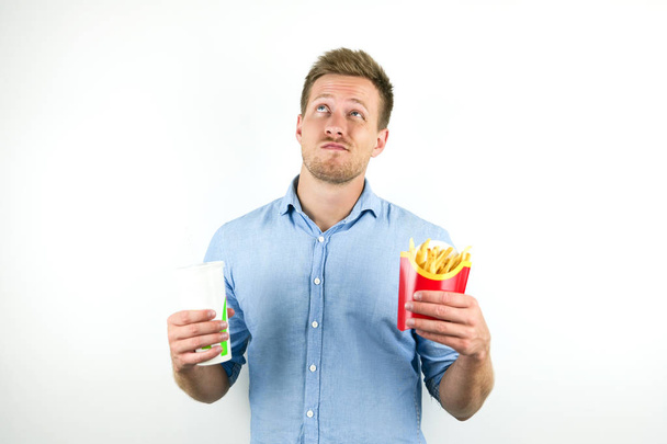 handsome young man holds soda in papper cup and fries from fast food restaurant looks doubtful on isolated white background - Foto, Bild