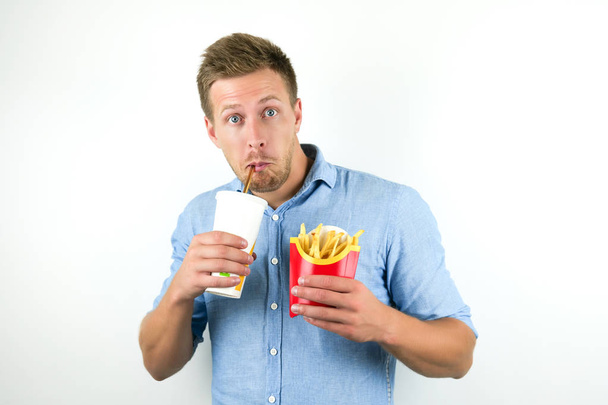joven guapo sostiene soda en taza de papper y patatas fritas de restaurante de comida rápida se ve sorprendido en el fondo blanco aislado
 - Foto, Imagen